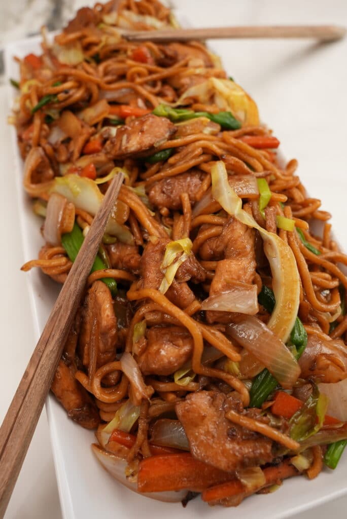 A close up photo of Chicken Lo mein on a rectangular tray with chopsticks.