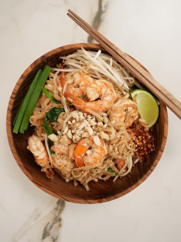 Pad Thai plated in a wooden bowl with lime, bean sprouts, and green onion.