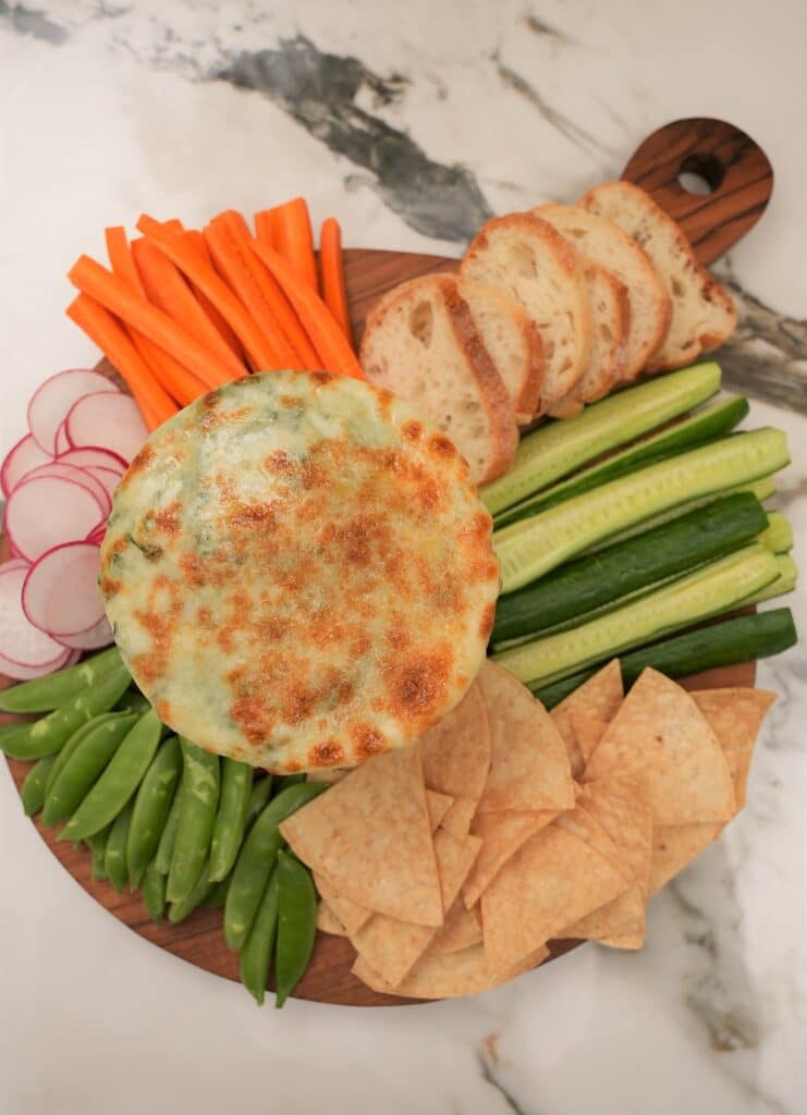 Spinach artichoke dip on a tray with carrots, vegetables, chips and bread.