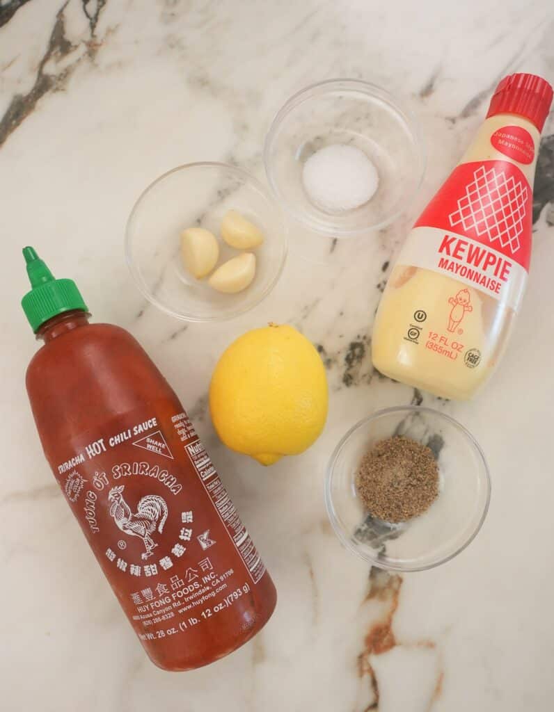 Spicy Garlic Mayo sauce ingredients on a table with garlic, salt, lemon, kewpie mayo, black pepper, and sriracha.