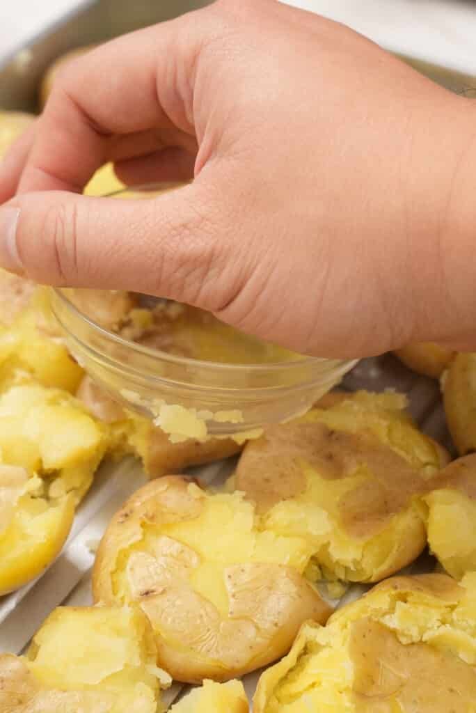 A hand smashing potatoes on a baking tray with a small bowl.