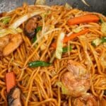 Shrimp Lo Mein cooked in a wok with shrimp, noodles, mushrooms, cabbage, carrots, scallions, and lo mein sauce.