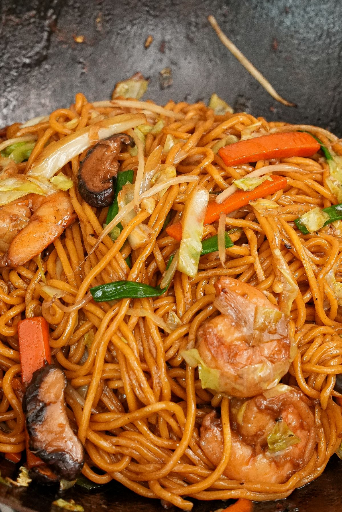 Shrimp Lo Mein cooked in a wok with shrimp, noodles, mushrooms, cabbage, carrots, scallions, and lo mein sauce.