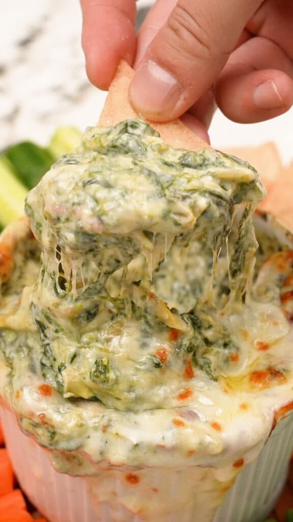 A hand pulling up Spinach Artichoke dip with a tortilla chip.