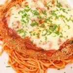 A plate of crispy chicken parmesan on a plate of spaghetti along with tomato sauce, parmesan cheese, mozzarella cheese and parsley.