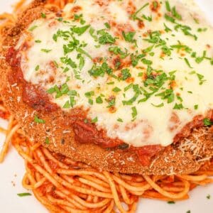 A plate of crispy chicken parmesan on a plate of spaghetti along with tomato sauce, parmesan cheese, mozzarella cheese and parsley.