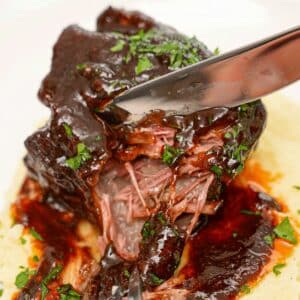 A tender braised short rib being pulled apart with a fork and knife on a bed of mashed potatoes.