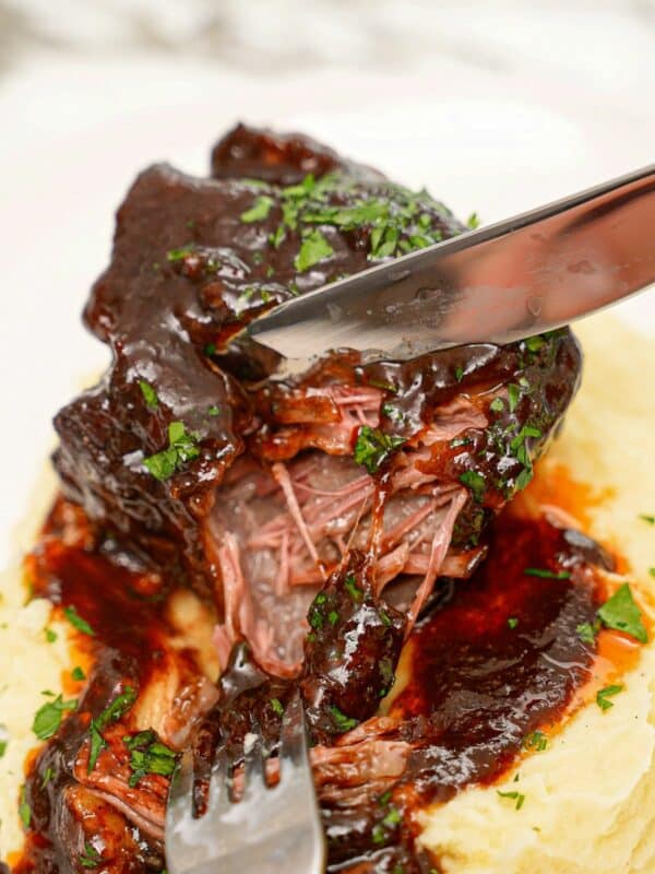 A tender braised short rib being pulled apart with a fork and knife on a bed of mashed potatoes.