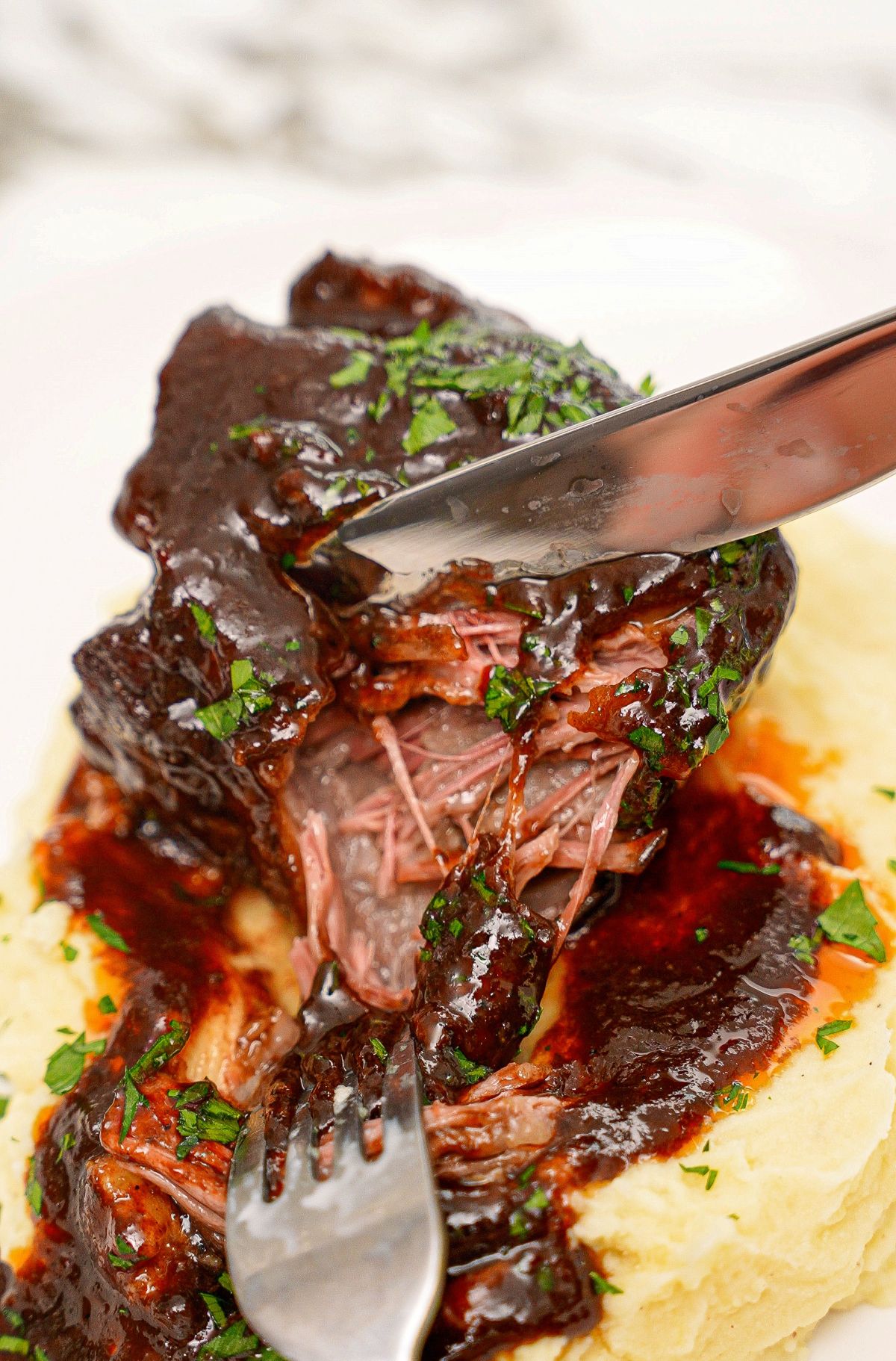A tender braised short rib being pulled apart with a fork and knife on a bed of mashed potatoes.