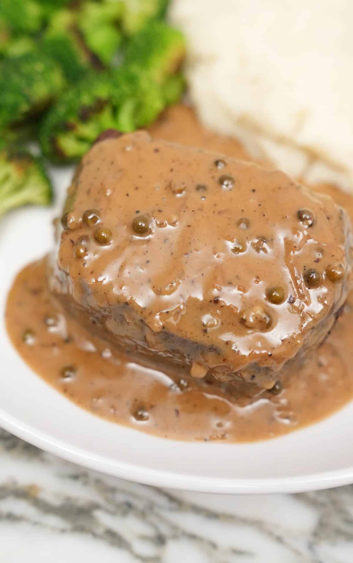 A plate of steak covered in a creamy peppercorn sauce next to broccoli and mashed potatoes.