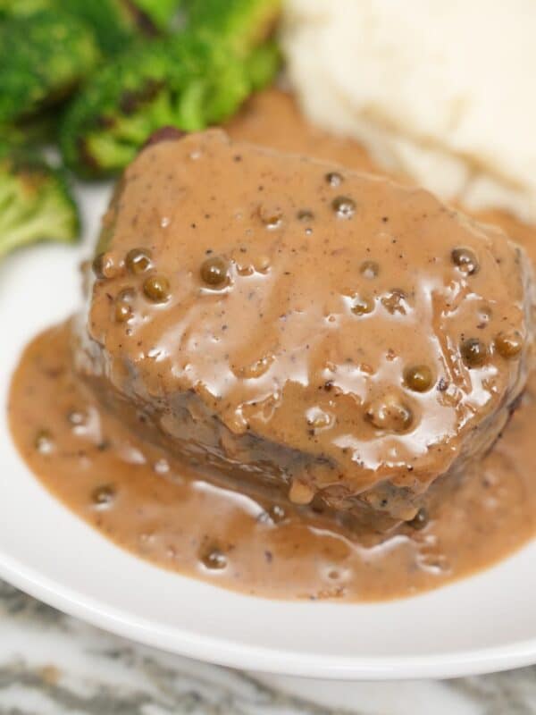 A plate of steak covered in a creamy peppercorn sauce next to broccoli and mashed potatoes.