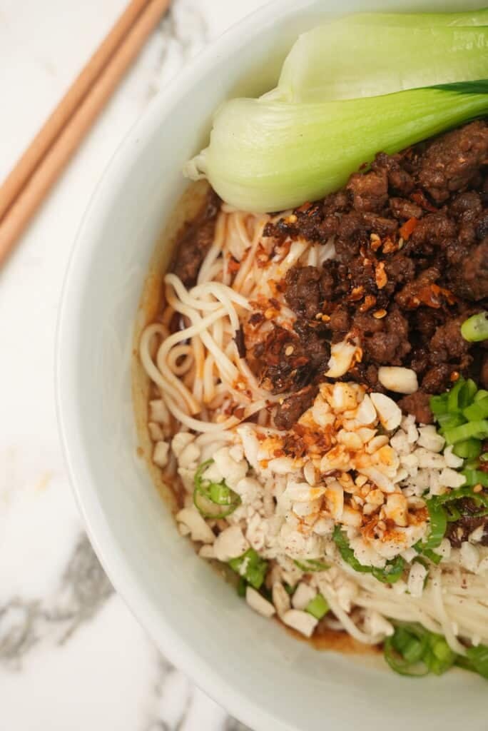 A close up photo of Dan Dan noodles in a bowl with bok choy and chopsticks.