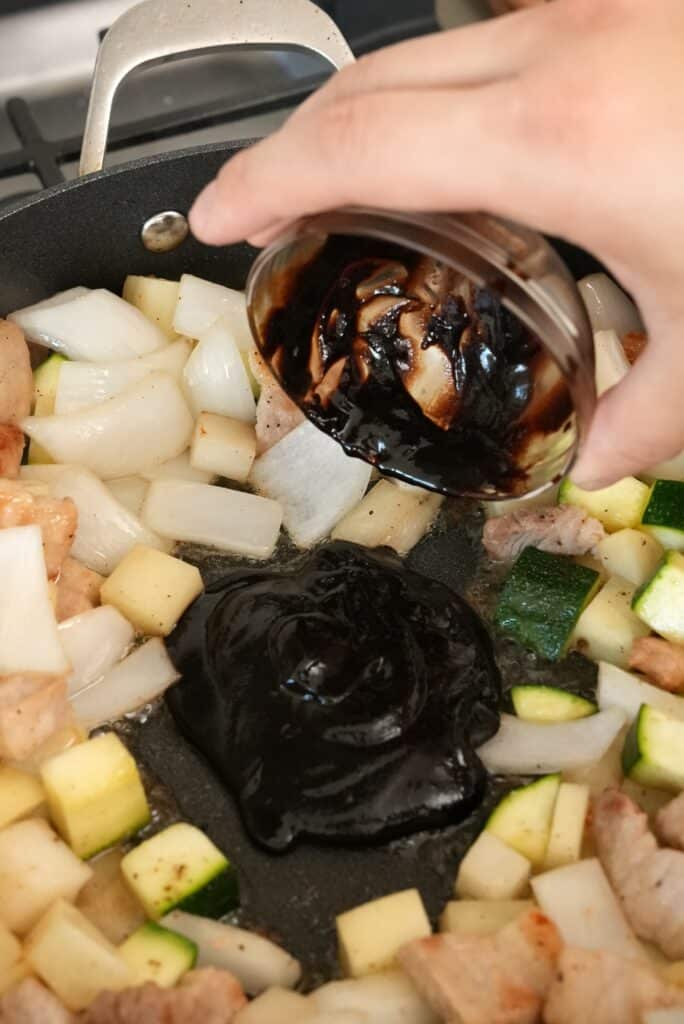 A hand adding Korean black bean paste to the center of the pan of pork belly and vegetables.