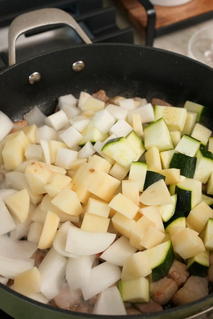 Potatoes, zucchini, radish, and onions being added to a pan with pork belly.