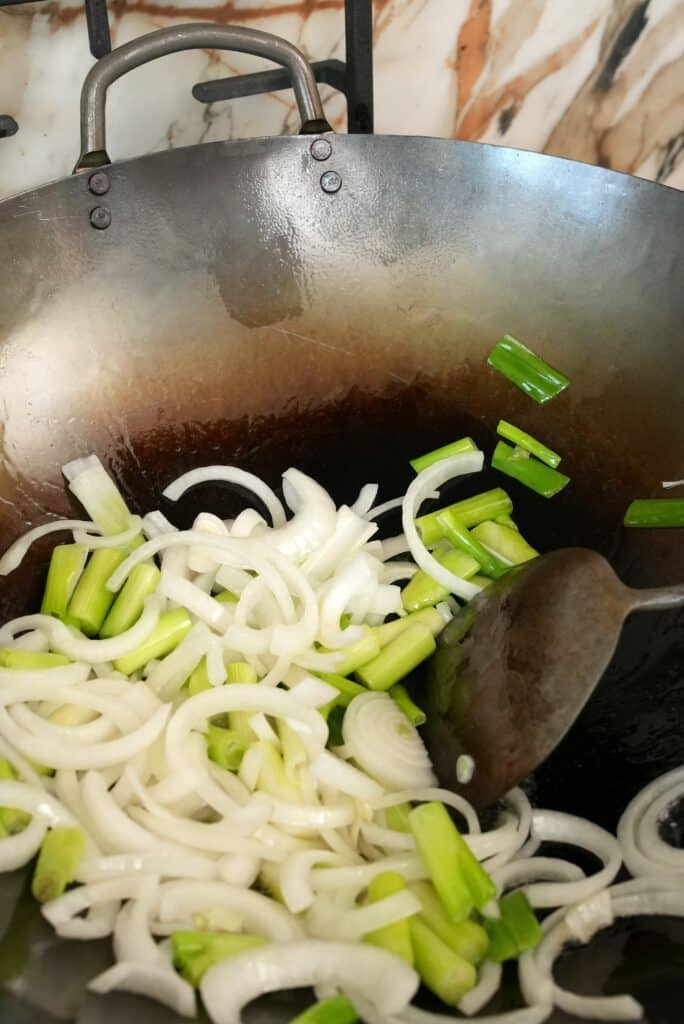Onions and green onion whites being stir fried in a wok with a spatula.