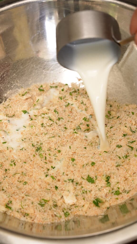 Whole milk being added to seasoned breadcrumbs in a metal mixing bowl.