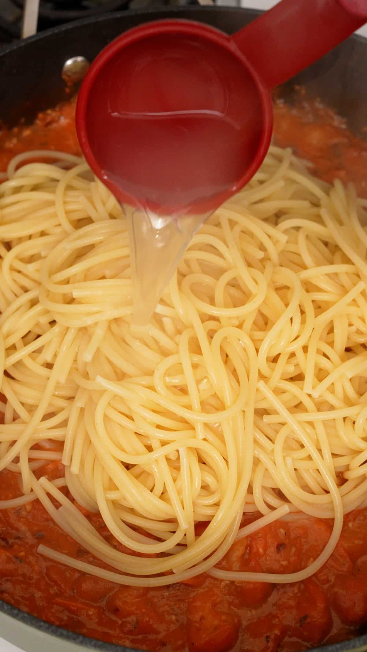 A measuring cup pouring pasta water into spaghetti and tomato sauce in a pan.
