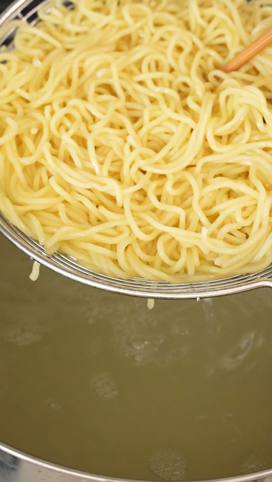 A metal spoon draining ramen noodles from boiling water.