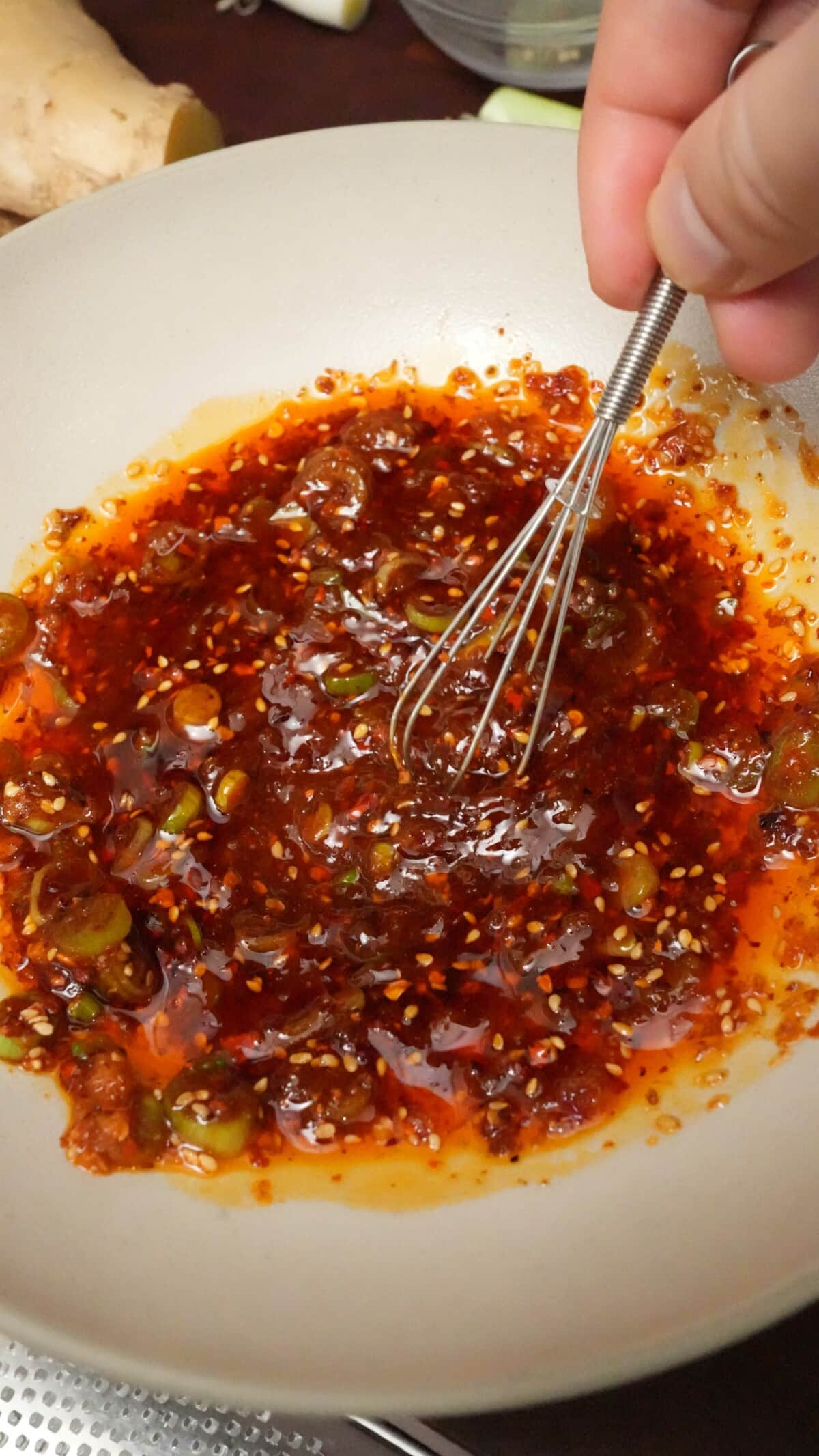 Hot oil being mixed with chili flakes and aromatics in a ceramic bowl with a small whisk.