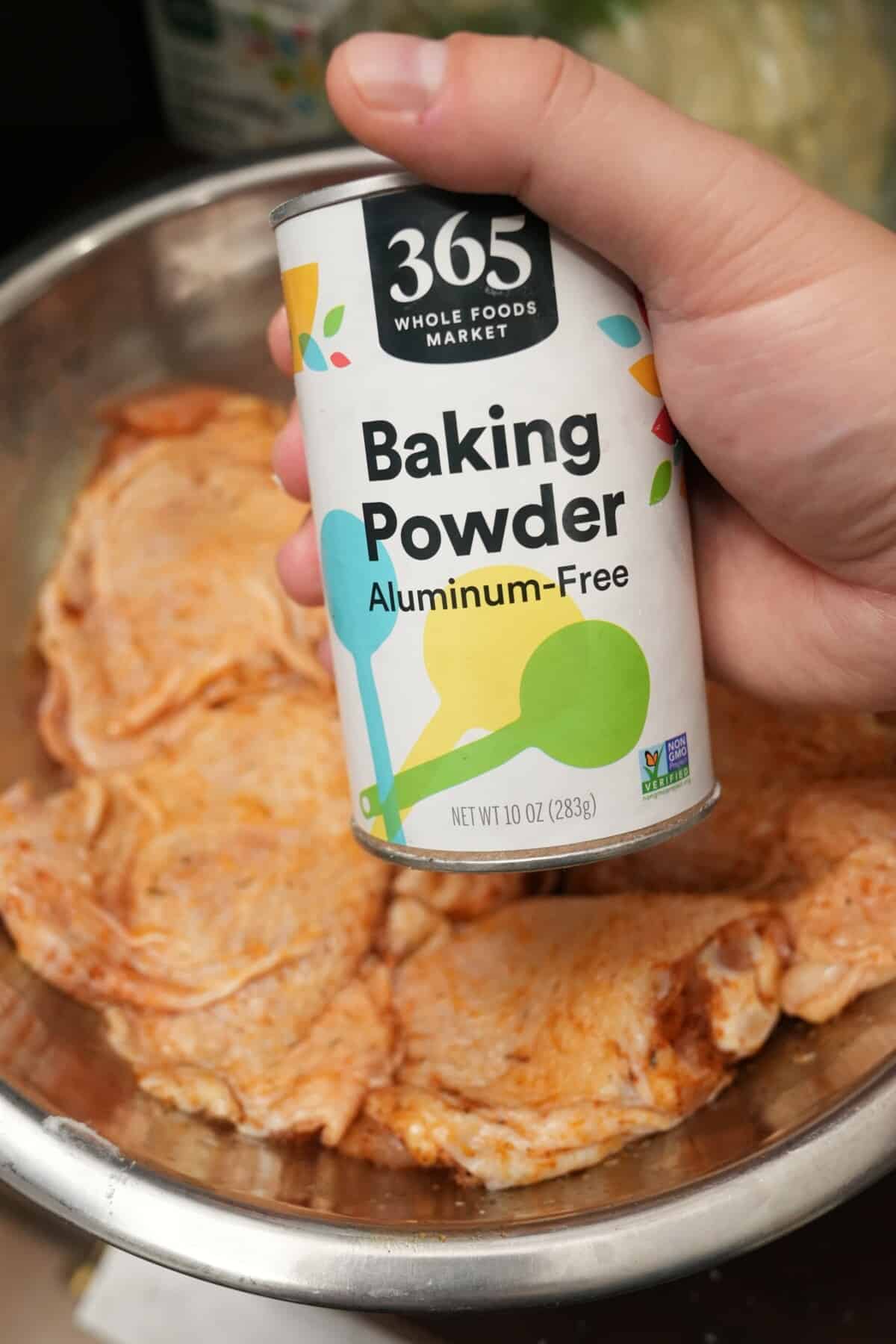A can of baking powder being held over a bowl of seasoned bone-in chicken thighs.