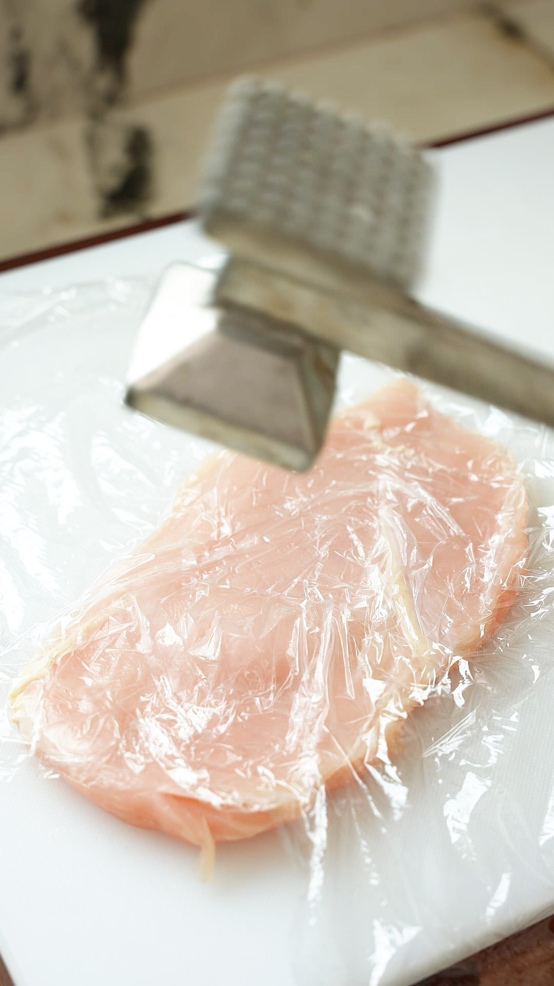 A meat mallet pounding chicken thinly on a cutting board.