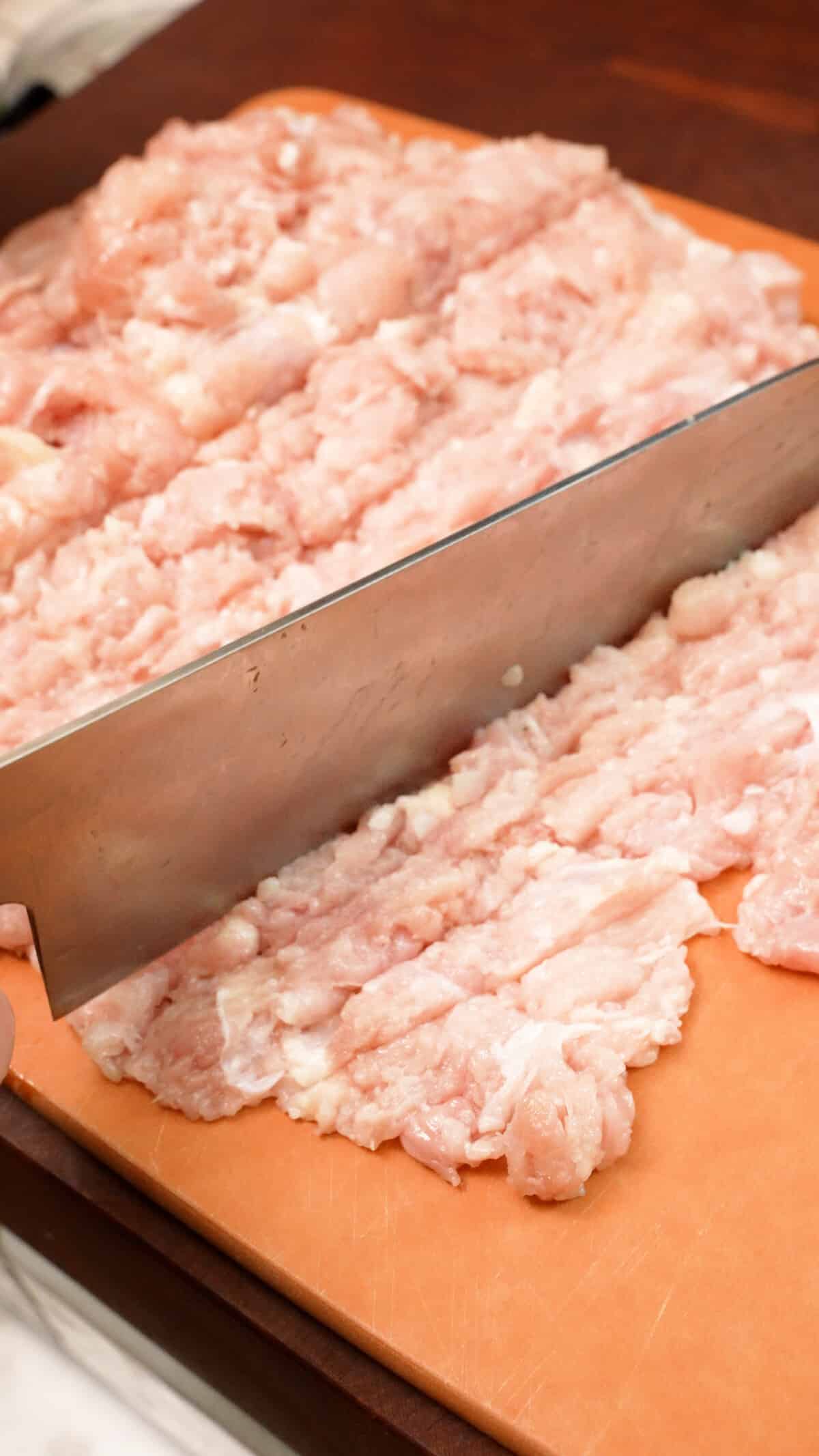 A knife mincing chicken thigh on a cutting board.