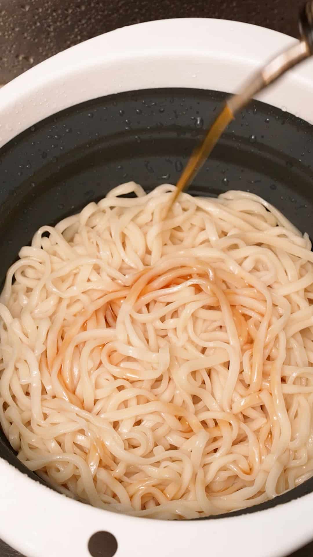 Adding sesame oil to the cooked, rinsed noodles in a strainer.