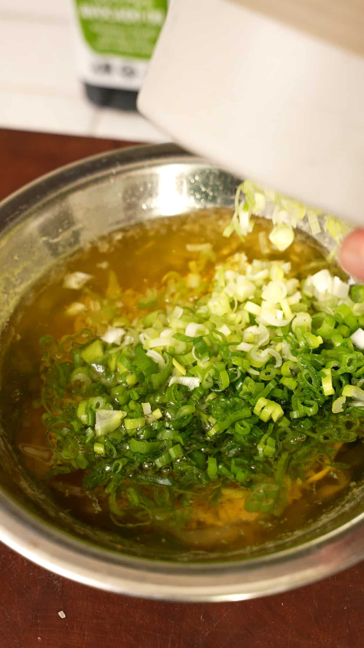 Scallions being added to a metal bowl with ginger oil.