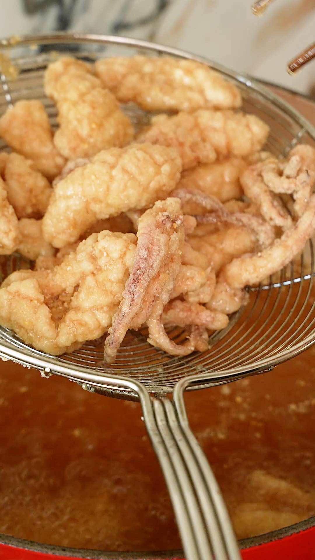 Freshly fried squid being drained from the frying oil by a mesh strainer.
