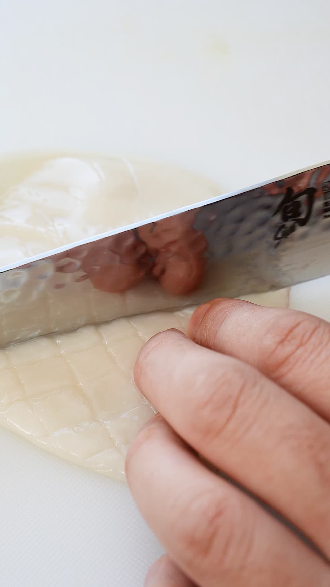 A squid tube being scored with a sharp knife on a cutting board.