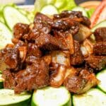 A plate of Shaking Beef (Bo Luc Lac) on a plate with cucumbers and tomatoes.