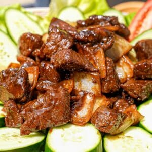 A plate of Shaking Beef (Bo Luc Lac) on a plate with cucumbers and tomatoes.