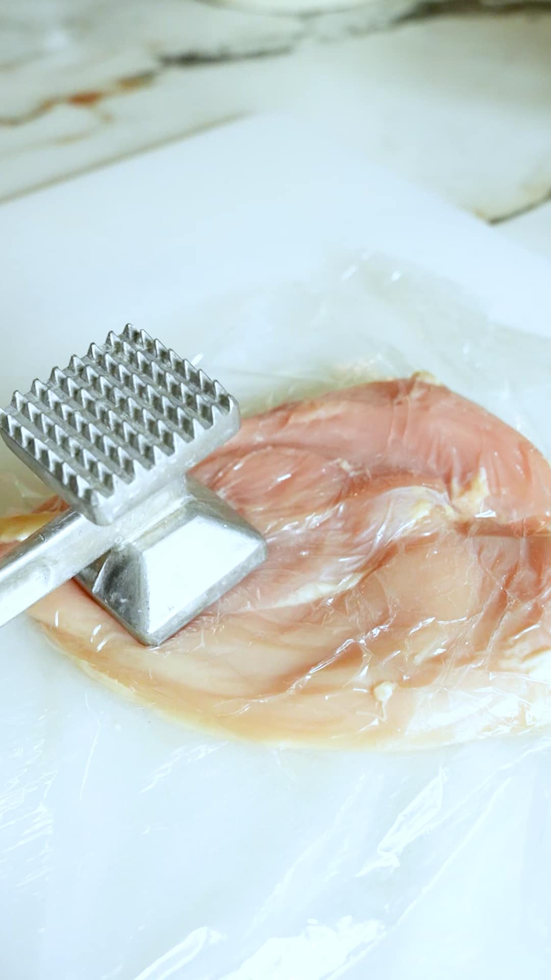 Chicken breast covered with plastic wrap being pounded thin on a cutting board with a meat mallet.