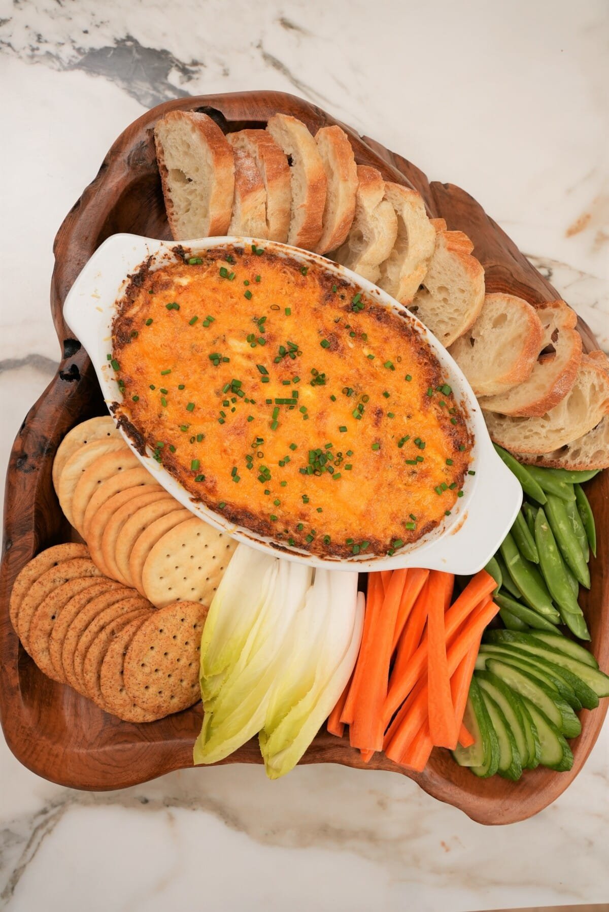 Baked crab dip on a wooden tray with bread, crackers, and vegetables.