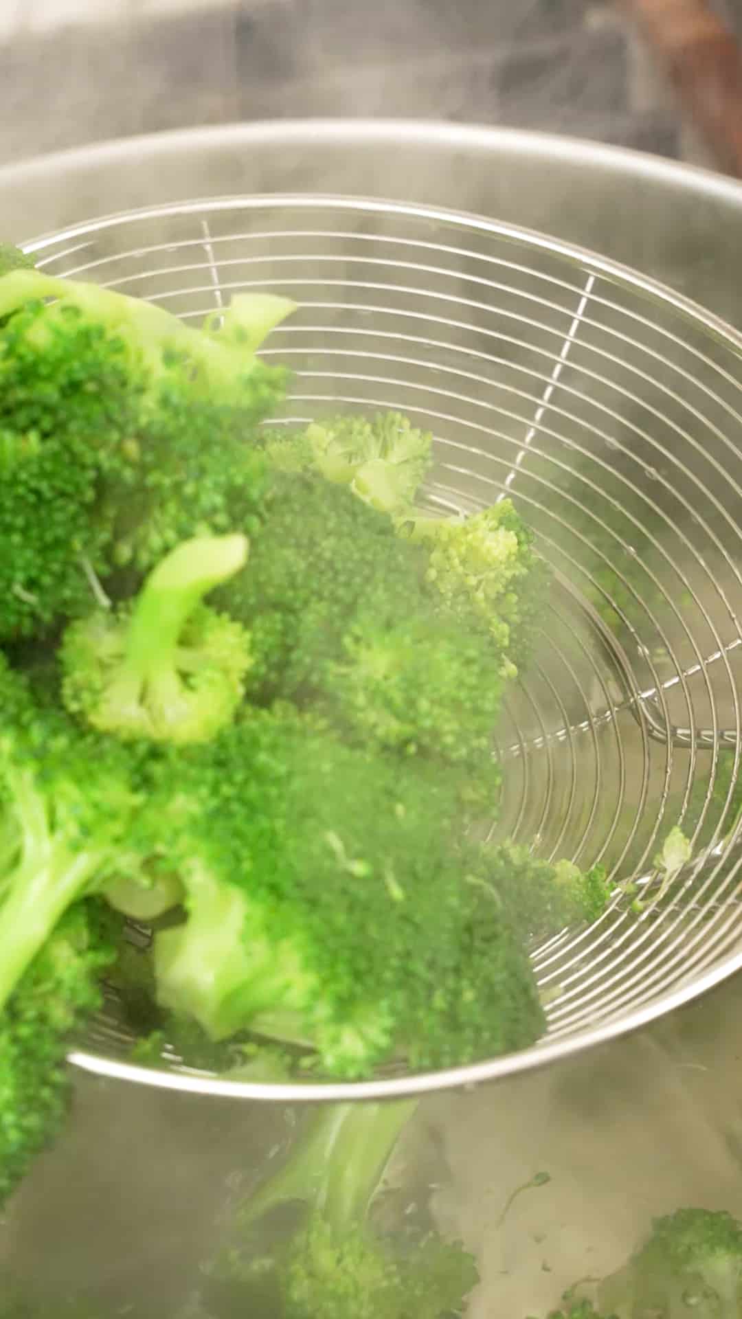 A metal strainer removing blanched broccoli from a pot of boiling water.