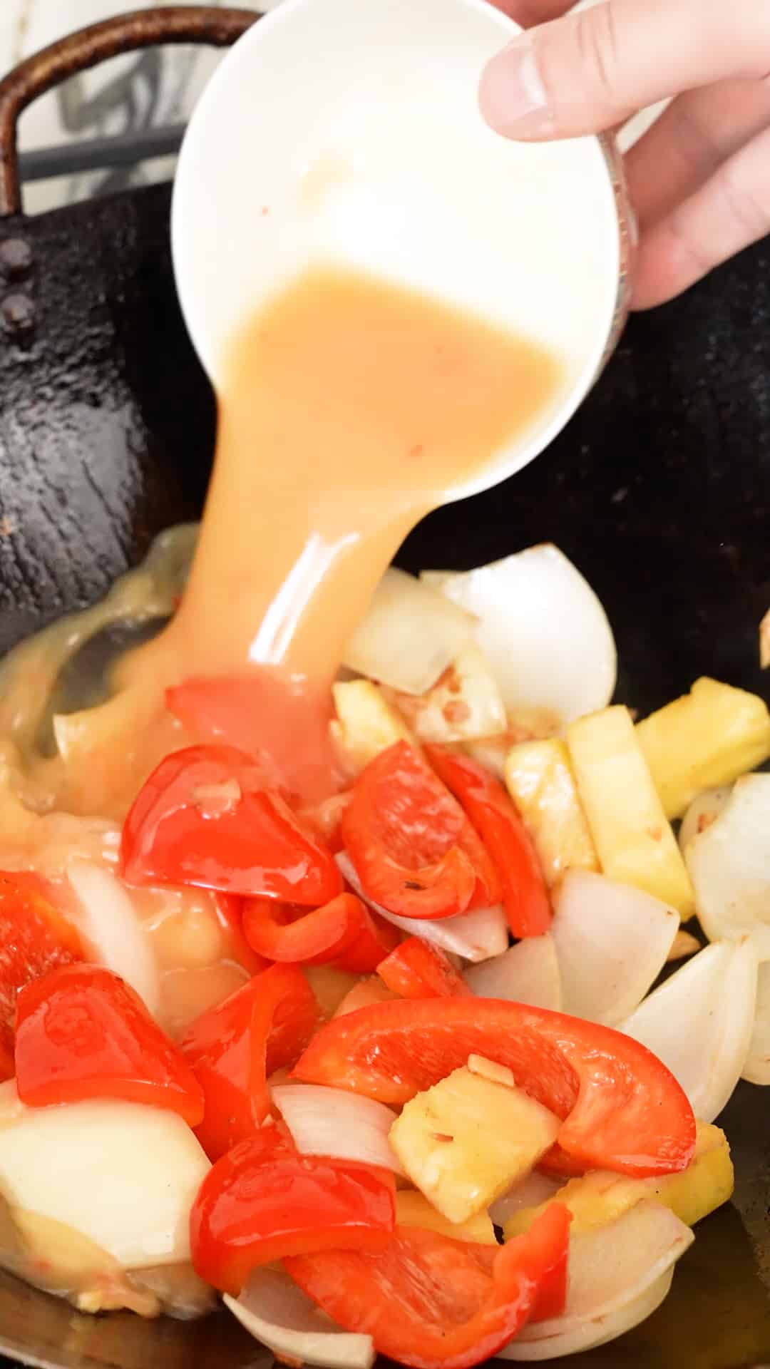 Sauce being added to the peppers, onions and pineapple in a wok.