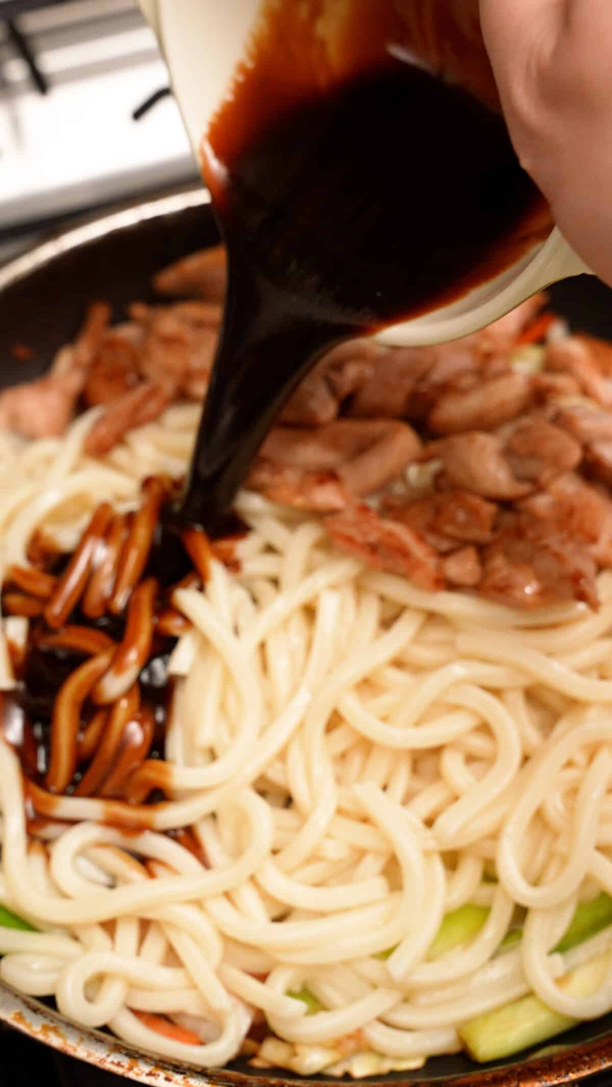 Yaki Udon stir fry sauce being added to noodles and chicken in a pan.