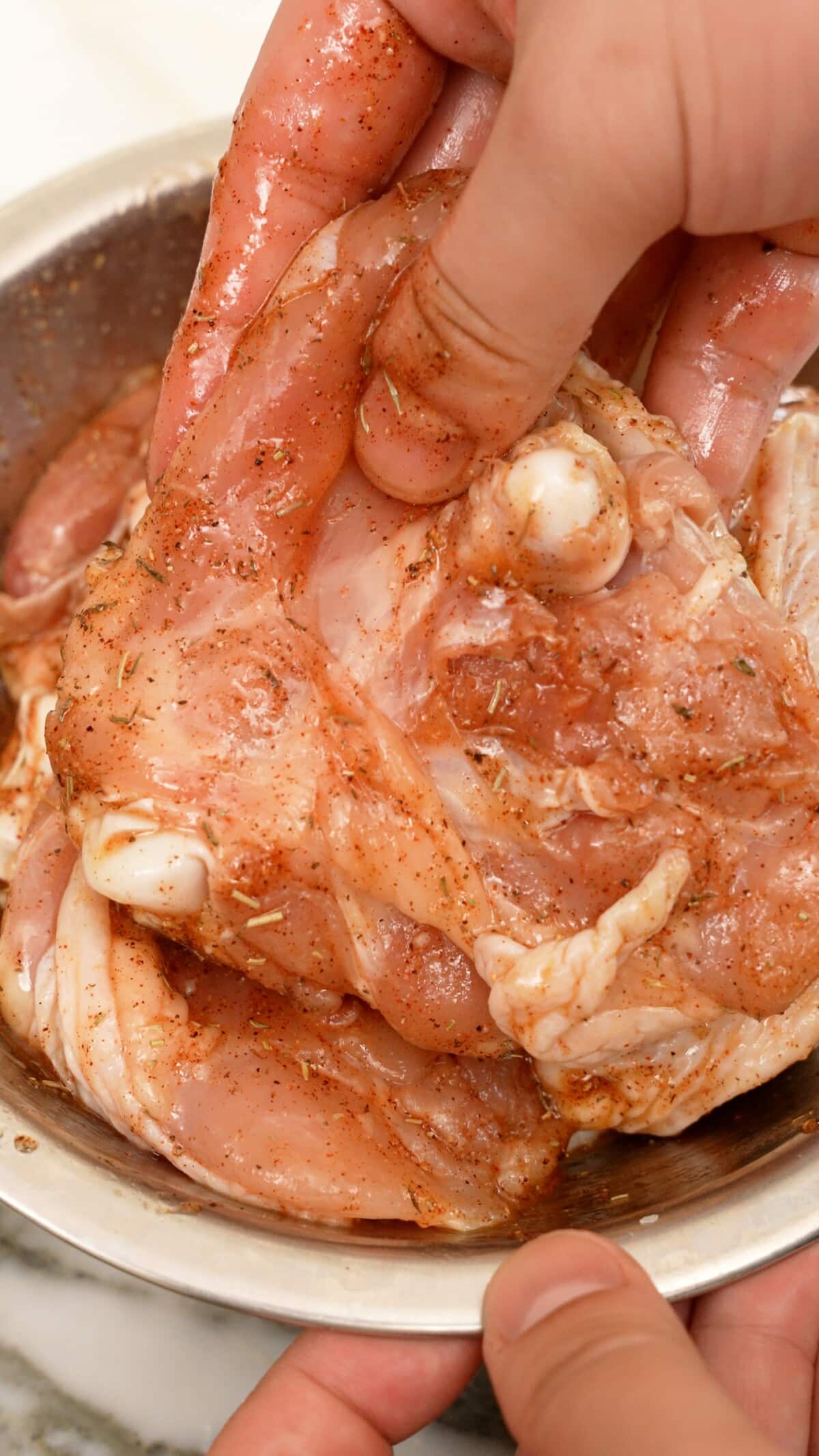 A hand seasoning holding a seasoned piece of chicken thigh in a metal bowl.