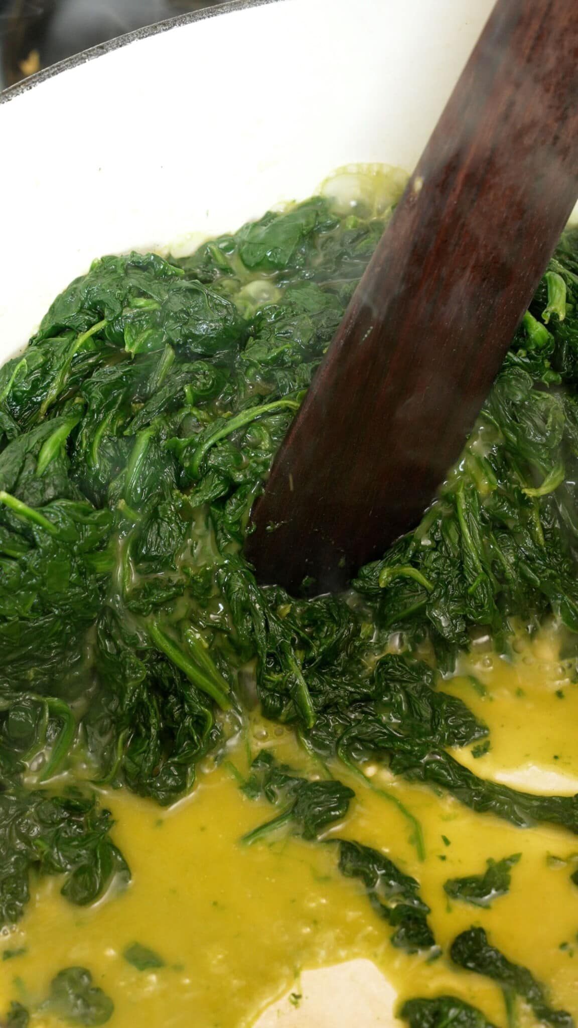 A wooden spatula cooking out the water from spinach in a pot.