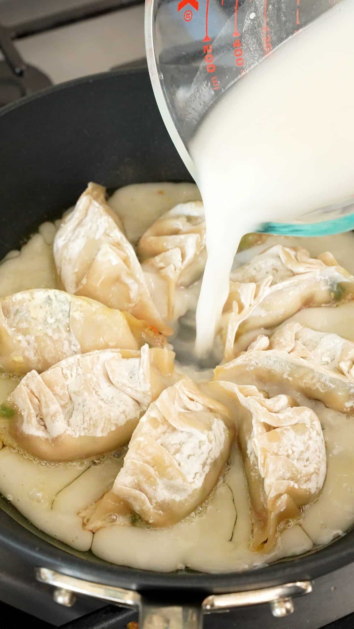 A measuring cup pouring dumpling skirt batter into the pan of dumplings.