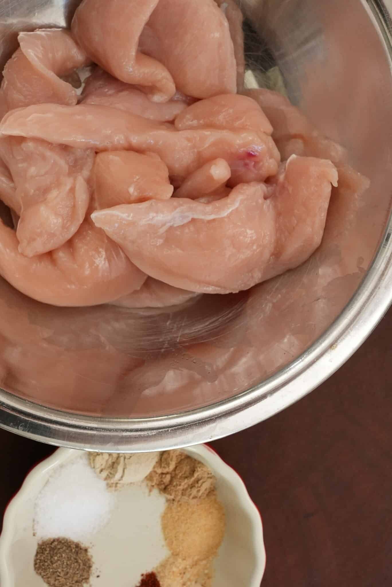 Raw chicken in a metal bowl next to a bowl of spices.