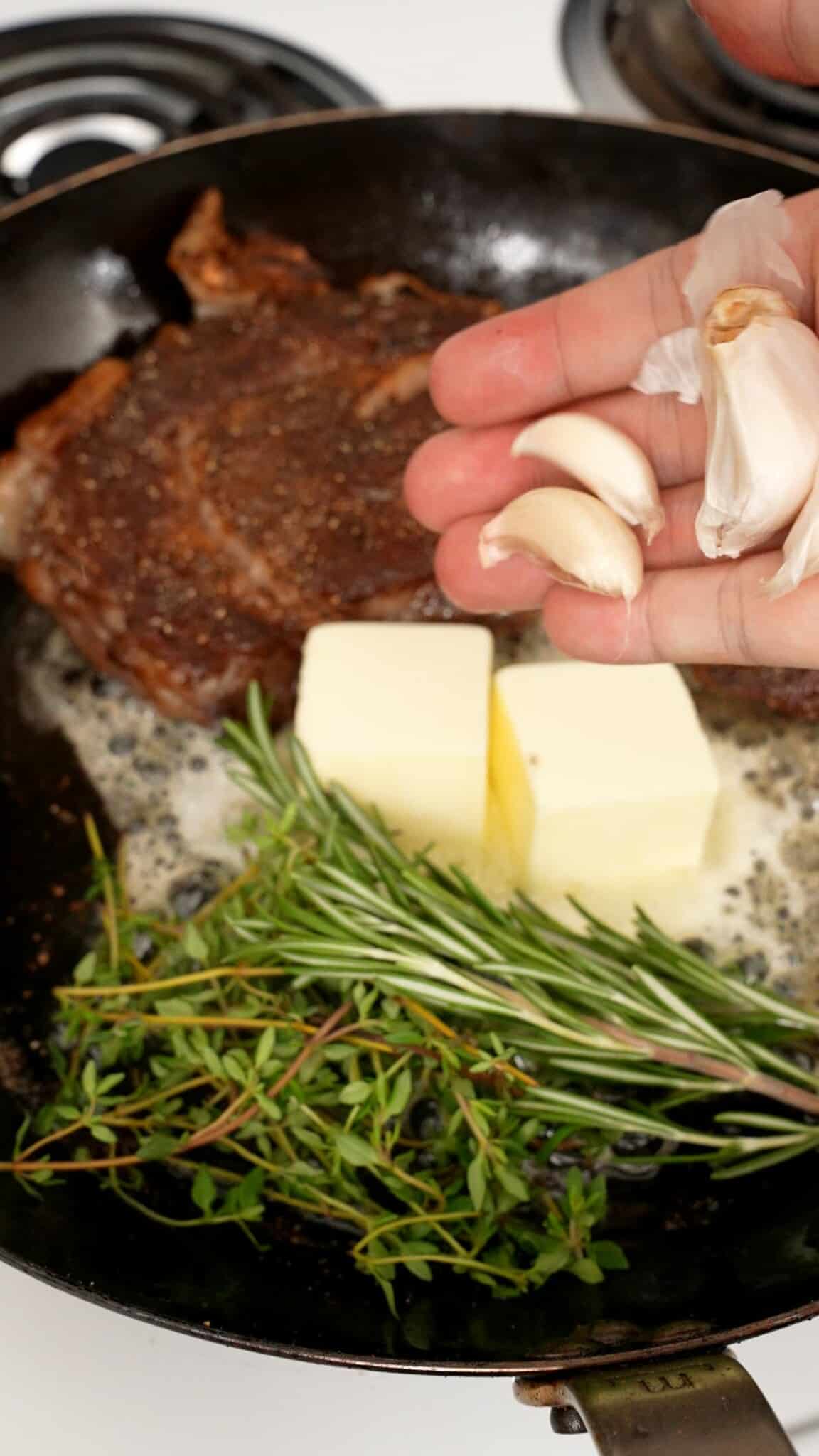 Raw garlic being added to a pan of butter and fresh herbs to baste a steak.