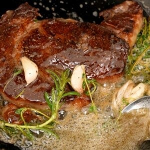 Steak searing in pan with fresh herbs and butter.