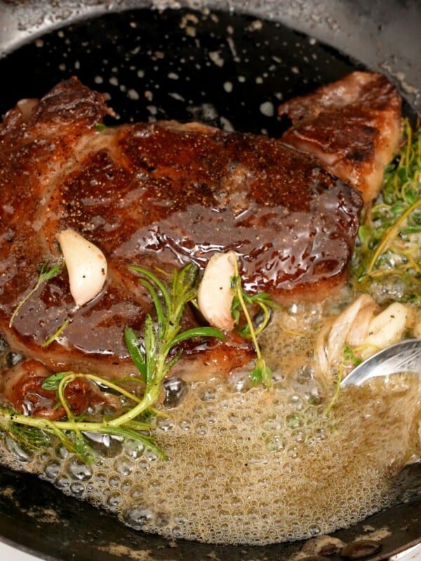 Steak searing in pan with fresh herbs and butter.