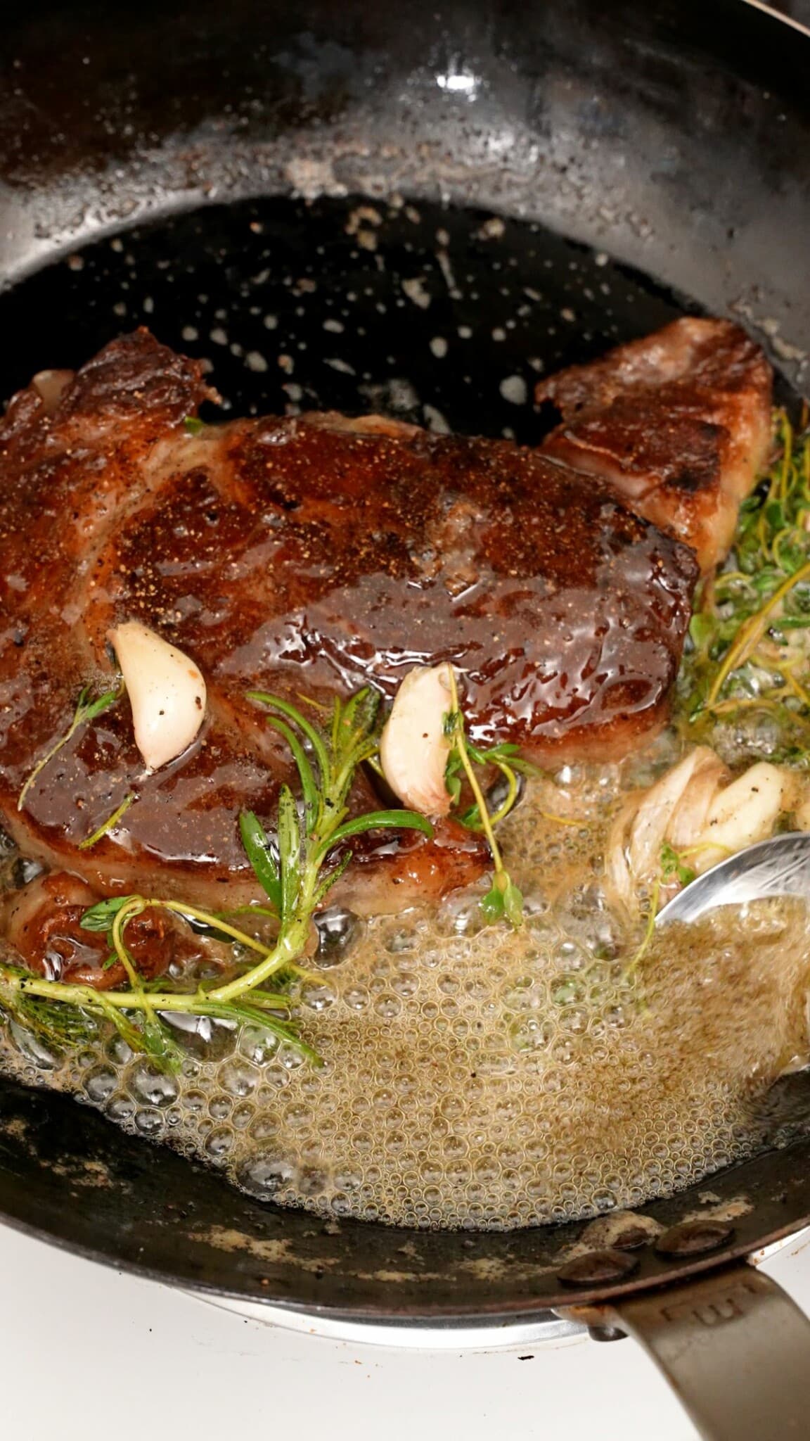 Steak searing in pan with fresh herbs and butter.