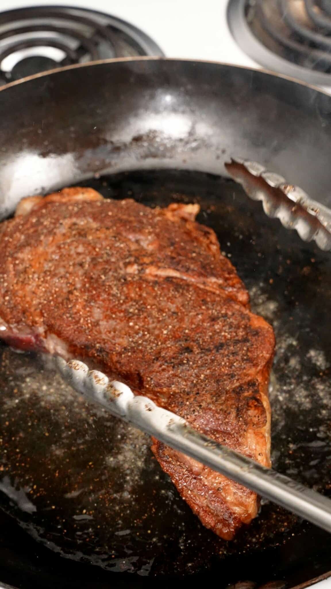 A pair of tongs searing the steak in a pan.