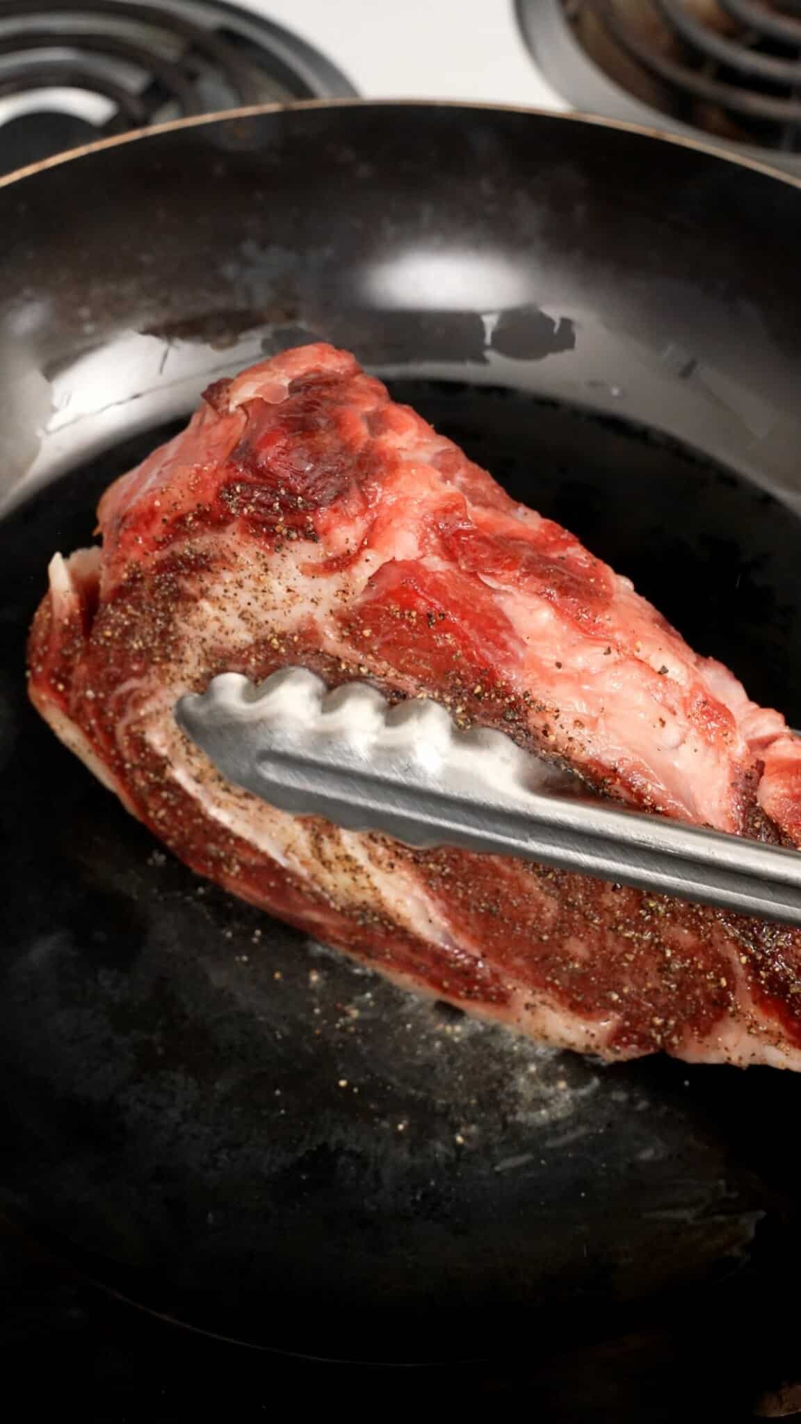 A pair of tongs searing the fat cap of the ribeye steak.