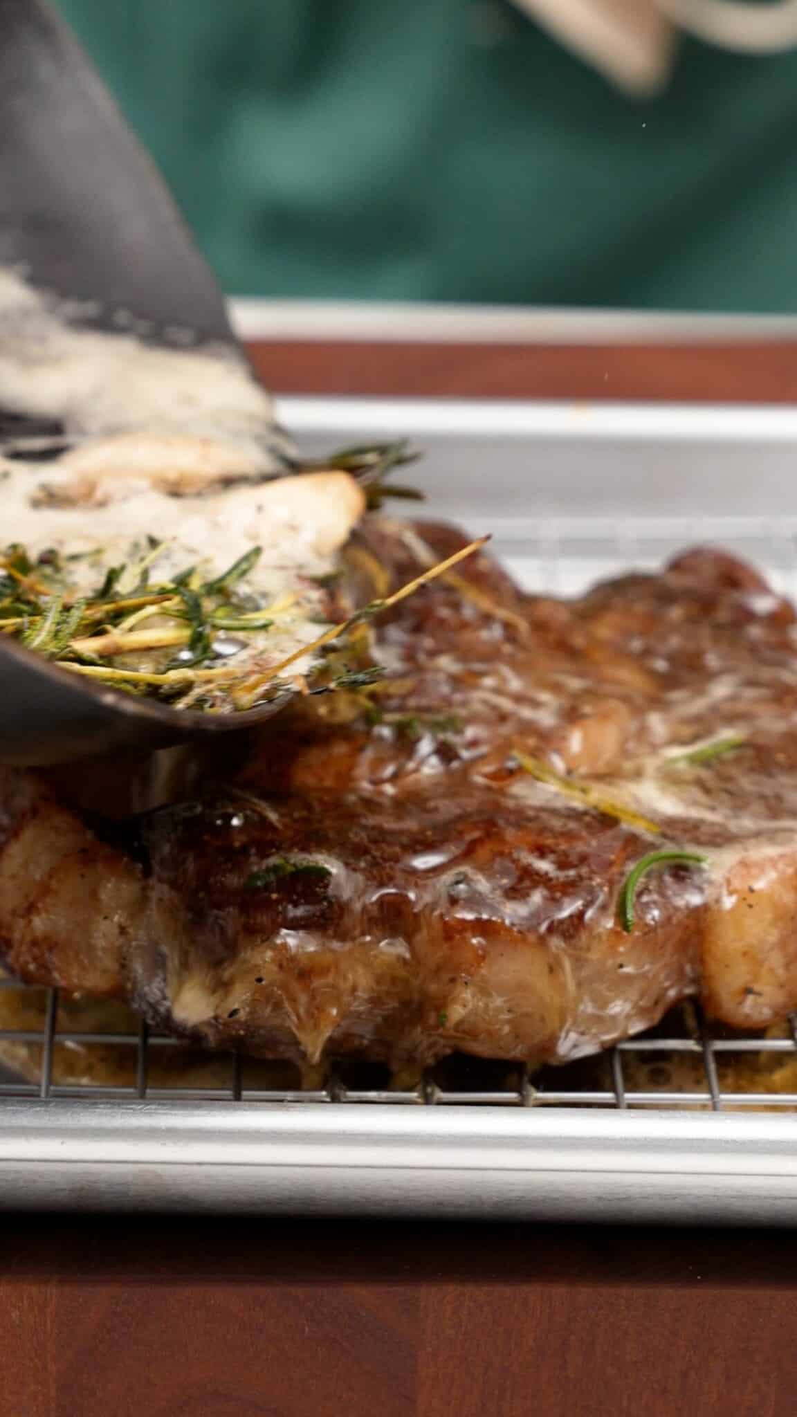 A pan pouring hot butter and herbs over a steak resting on a wire rack.