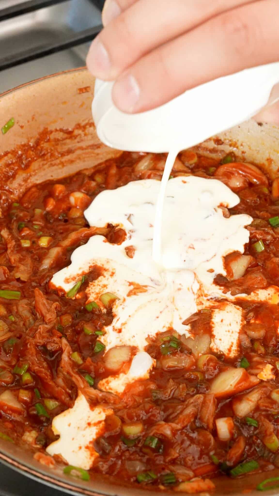 Adding heavy cream in a pan with scallions, kimchi and gochujang sauce.