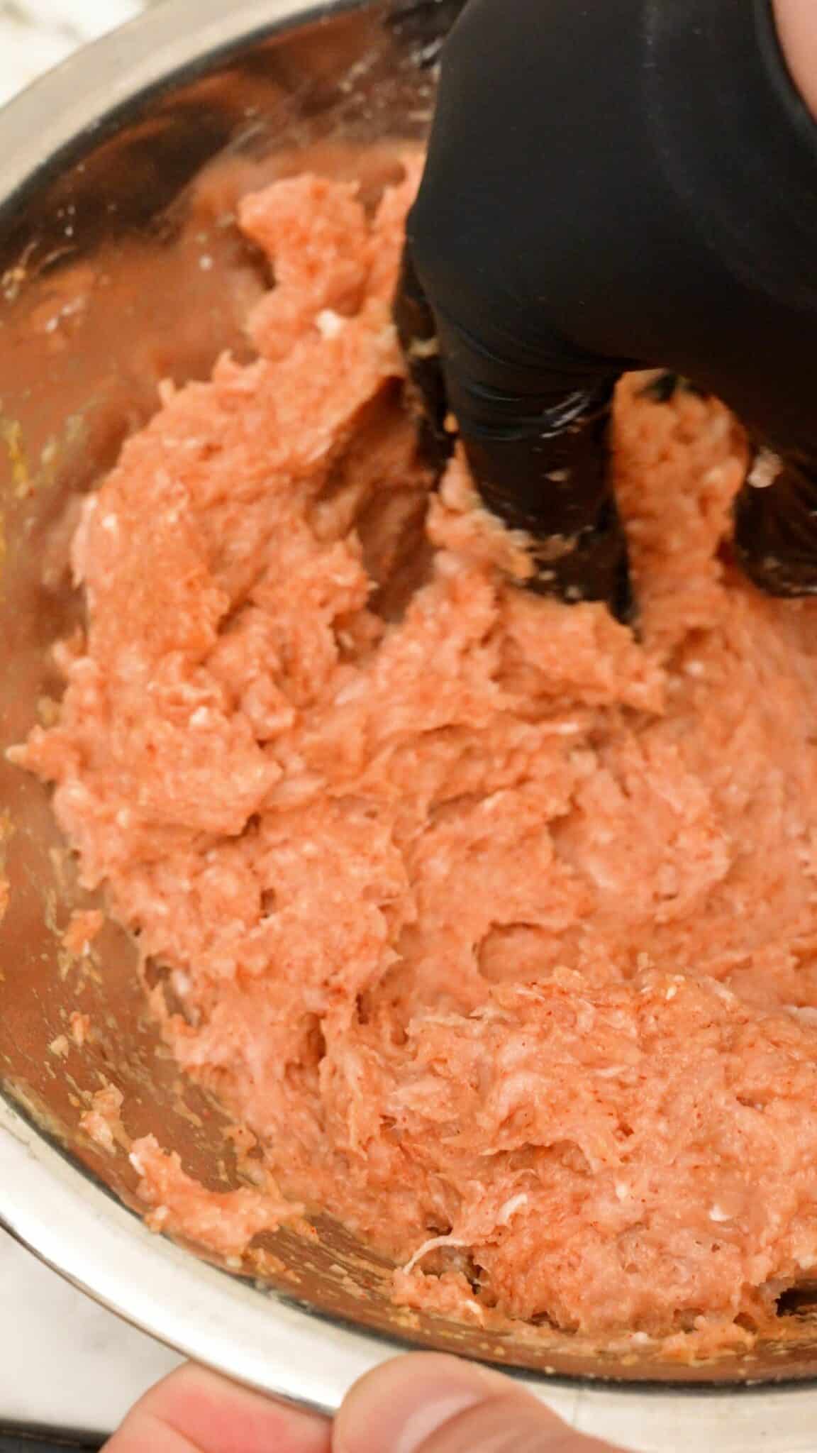 A hand mixing ground chicken, egg, cornstarch, and spices in a bowl.